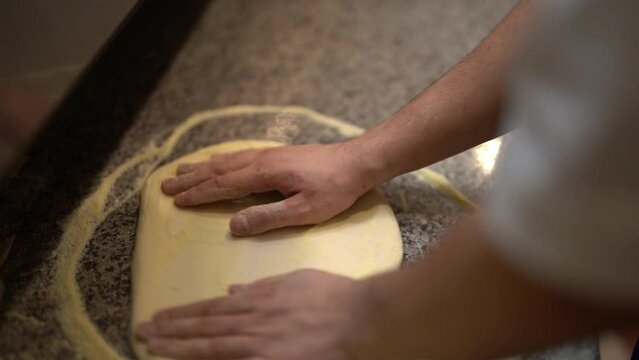 Side crop of unanimous male pizza chef kneading and rotating dough while making in front of grey marble cooking table in pizzeria's kitchen 