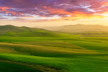 Obraz premium beautiful green valley with green fields with green spring grass with nive hills and mountains and scrnic colorful cloudy sunset on background of landscape