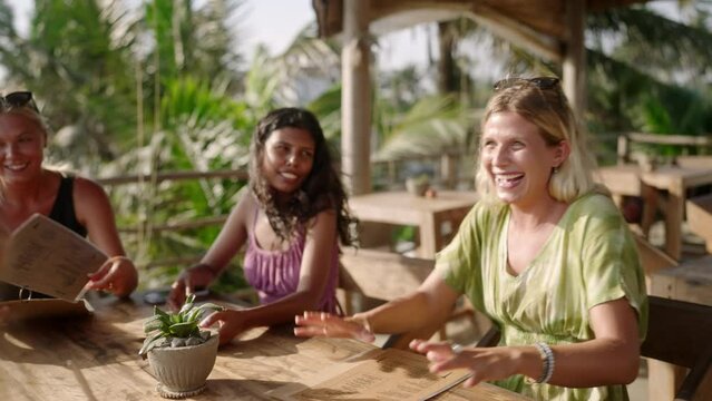 Diverse female friends in outdoor restaurant chatting. Multiracial cheerful girls sitting at the table in a tropical cafe talking to each other choosing food. Young multiethnic women having dinner