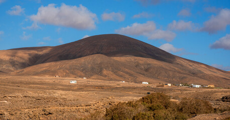 Vista panorámica de un paisaje desértico y volcánico con una gran montaña rocosa en la parte trasera y debajo de un pequeño pueblo con típicas casas blancas de Fuerteventura en Islas Canarias
