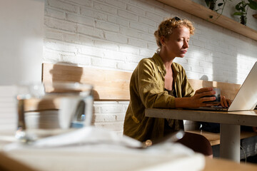 A young girl works in a cafe at a laptop, drinking hot coffee.