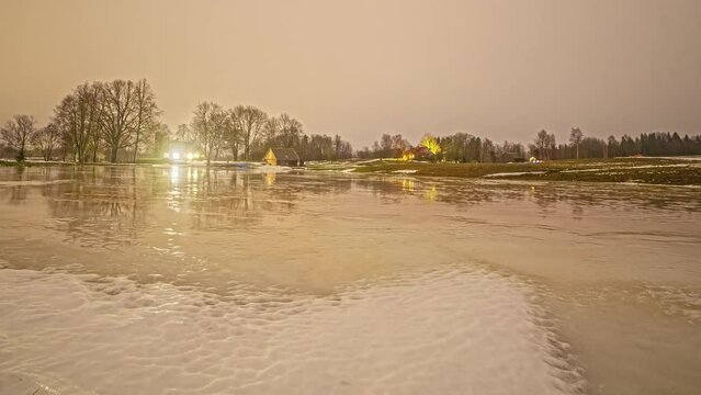 Timelapse shot of melting snow in front of cottages along rural countryside at night time.