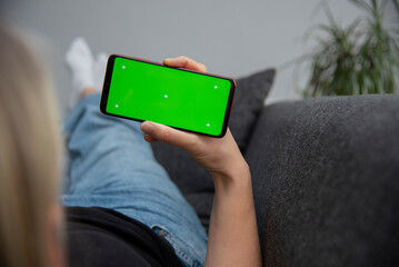 Back view of young europen woman who is using chroma key smartphone screen.She is sitting on the sofa in her cozy living room.