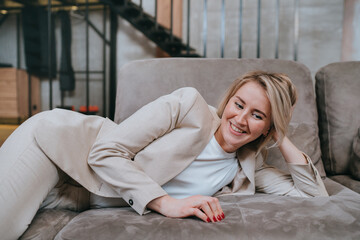 Fototapeta premium Relaxed short haired blonde young woman laying on couch smiles wearing beige business suit enjoys new furniture home. Successful businesswoman happy after meeting. Female dreaming.