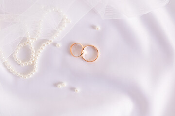 Two gold wedding rings on white satin textiles with pearl beads and veil . Wedding festive background. A copy of the space.