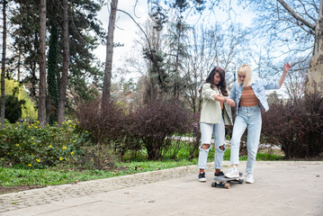 Two cool-looking hipster friends in park learning to ride a skateboard, having fun between studying or working, spending time together in a nice sunny weather. A friend teaches her roommate to skate