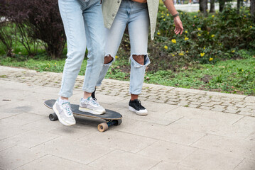 Two cool-looking hipster friends in park learning to ride a skateboard, having fun between studying or working, spending time together in a nice sunny weather. A friend teaches her roommate to skate