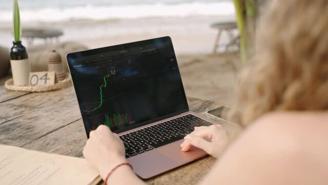 Female cryptocurrency trader at laptop checking candlestick charts online working remotely at outdoor tropical seaside cafe. Woman crypto broker analyses stock exchange rates and bets by ocean closeup