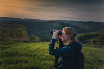Obraz premium Portrait of a young female tourist looking through binoculars at sunset while walking on green summer mountain valley