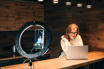 Social media influencer creating content at modern studio sharing online stream using smartphone and laptop. Girl blogger smiling and speaking recording video  with ring lamp. Online training courses.