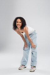 full length of carefree african american woman in blue jeans smiling at camera on grey background.