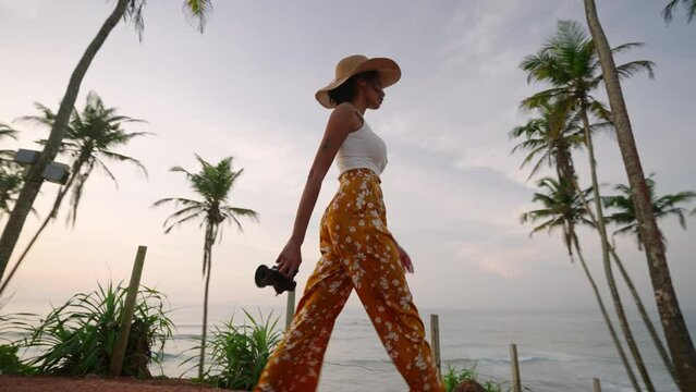 African american woman walking with camera on tropical island. Female multiracial photographer tourist explores ocean view location on vacation. Black girl in straw hat taking pictures on sunrise.