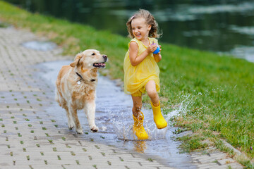 A child in a yellow dress and rubber boots along with her dog, jumps through puddles after the rain.
