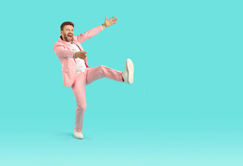 Full body portrait of a cool casual young funny guy with unshaven beard dancing wearing bright pink suit isolated on blue turquoise background. Happy excited man having fun in studio.