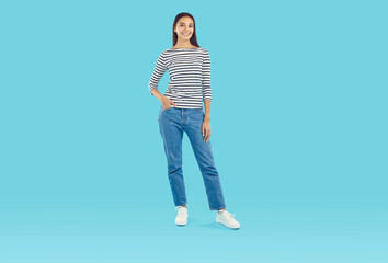 Naklejka premium Full body shot of a beautiful young Caucasian woman wearing a comfortable striped three quarter sleeve top and blue straight leg jeans standing on a blue studio background. Casual fashion concept