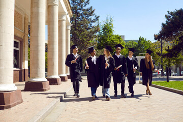 Happy smiling university students and friends in graduate caps and gowns talk and share plans for future while walking in beautiful campus yard after outdoor graduation ceremony on sunny summer day