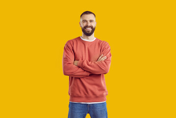 Young smiling bearded man in red sweatshirt and jeans is posing with arms crossed on chest standing isolated on yellow background looking at camera. Sincere human emotions, body language concept. - obrazy, fototapety, plakaty