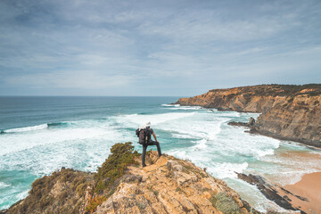 Hiker and adventurer stand on a rugged rock and cliff coastline and enjoy the view on the Atlantic Ocean in the west of Portugal in the famous tourist region of the Algarve