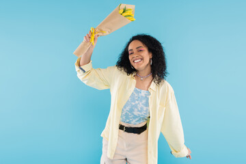 joyful and trendy african american woman smiling at camera while holding yellow tulips isolated on blue.