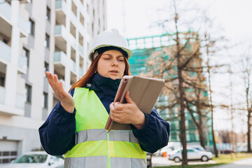 Architect at a construction site. Portrait of woman constructor wearing white helmet and safety yellow vest. Upset sad, skeptical, serious woman looking at the tablet screen outdoors.