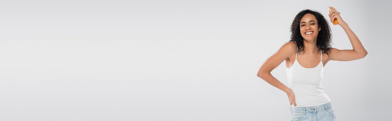 smiling african american woman standing with hand on hip and spraying hair oil isolated on grey, banner.