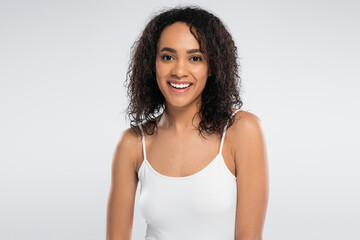 portrait of carefree african american woman in white tank top smiling at camera isolated on grey.