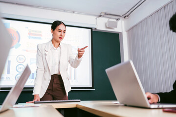 A cheerful and confident Asian businesswoman stands, present bar charts data from projector screen to her office colleagues. Asian business women leader role at the meeting.