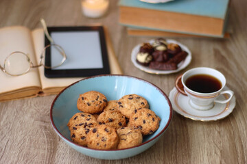 Books, reading glasses, e-reader, plate of chocolate pralines, bowl of cookies, cups of tea and lit candles on the table. Selective focus.
