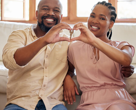 Young African American Couple Smiling Making A Heart Gesture With Their Hands Holding A Key Sitting On The Floor In Their New House Together. Husband And Wife Happy To Be Moving Into Their Own Home