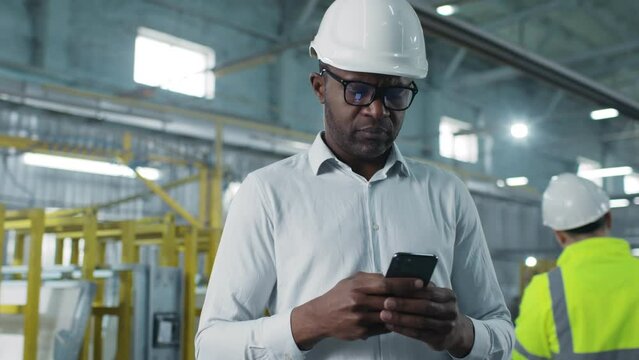 Careful concerned hard-working African-American male wearing white helmet marvelous glasses and white shirt looking around. Man using his superb gadget mobile phone.
