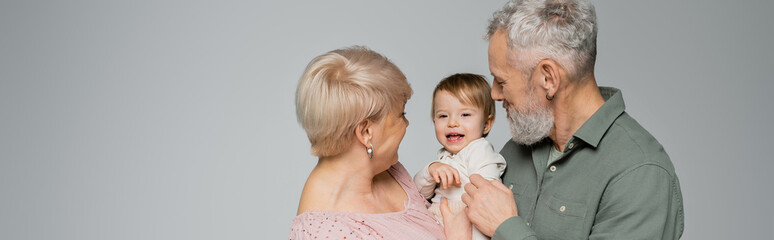 mature woman with bearded husband looking at cheerful toddler granddaughter isolated on grey, banner.