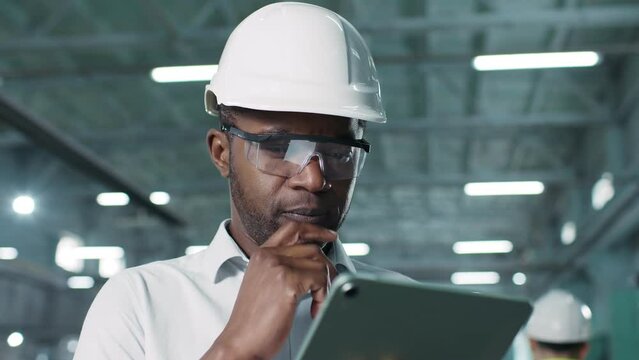Close-up portrait of thoughtful cautious mindful attentive African-American hard-working adult male. Unshaved man in hard hat typing in tablet tapping screen nodding his head.