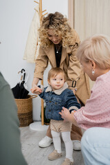 smiling woman in trench coat and toddler girl in denim jacket visiting grandparents at home.
