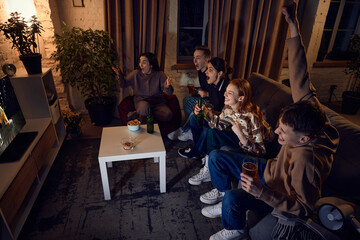 Group of young, emotional friends sitting on couch at home in the evening and watching live translation of basketball game. Concept of friendship, leisure activity, weekends, fun, emotions