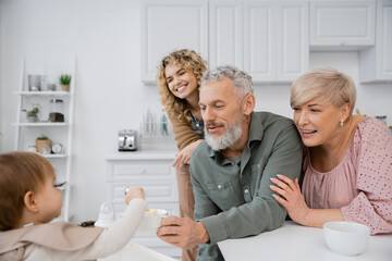 Obraz premium smiling bearded man holding bowl while feeding kid with breakfast near family in kitchen.