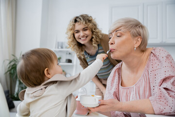 Obraz na płótnie Canvas little girl with spoon feeding grandma near smiling mother in kitchen.