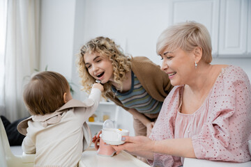Obraz na płótnie Canvas toddler girl feeding mom near smiling grandmother with bowl in kitchen.