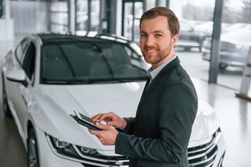 Standing, side view, holding tablet. Man in black suit is indoors in the car dealership near automobile