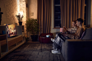 Young beautiful couple, man and woman spending time together at home in evening, sitting on couch, watching tv and eating popcorn. Concept of relationship, leisure activity, weekends, fun, emotions