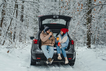 a couple sits in the trunk of their car and drinks coffee in a snowy winter forest