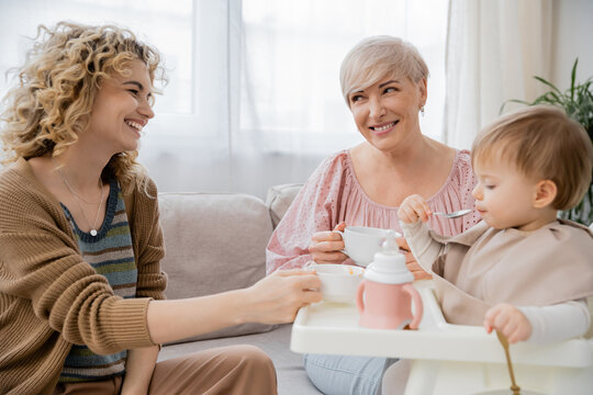 happy mature woman with tea cup looking at adult daughter feeding toddler girl in kitchen.