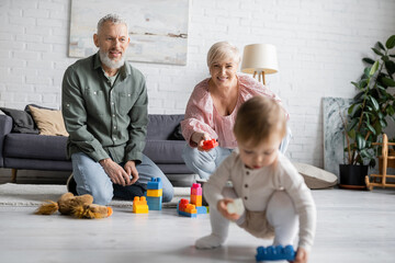 happy middle aged grandparents sitting on floor near building blocks while little girl playing with toy car in living room.