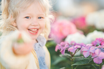 Fototapeta premium Closeup of a little blonde girl in blue shirt holding a flower in one hand with cute expression against the background of hydrangea flowers. Happy childhood