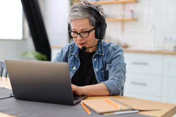 Focused mature woman works in headphones with a microphone on a laptop.