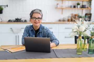 Senior woman at workplace in home interior, smiling and typing on laptop.