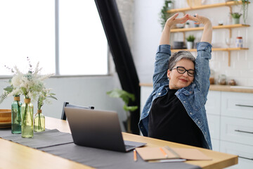 Positive senior woman doing stretching at the workplace in front of a laptop.