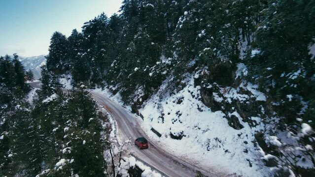 Aerial view of beautiful hilly roads covered with snow during winters in the Himalayan region of Uttarakhand. Snow-covered trees and cars moving on steep mountain roads. Drone following a car in hills