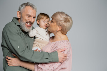 Fototapeta na wymiar mature woman kissing granddaughter near pleased bearded husband isolated on grey.