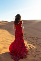 Young beautiful woman in long red dress with red rose petals among the desert. Desert rose...