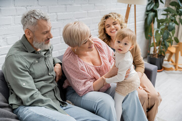 baby girl looking at camera near cheerful mother and grandparents on couch in living room.